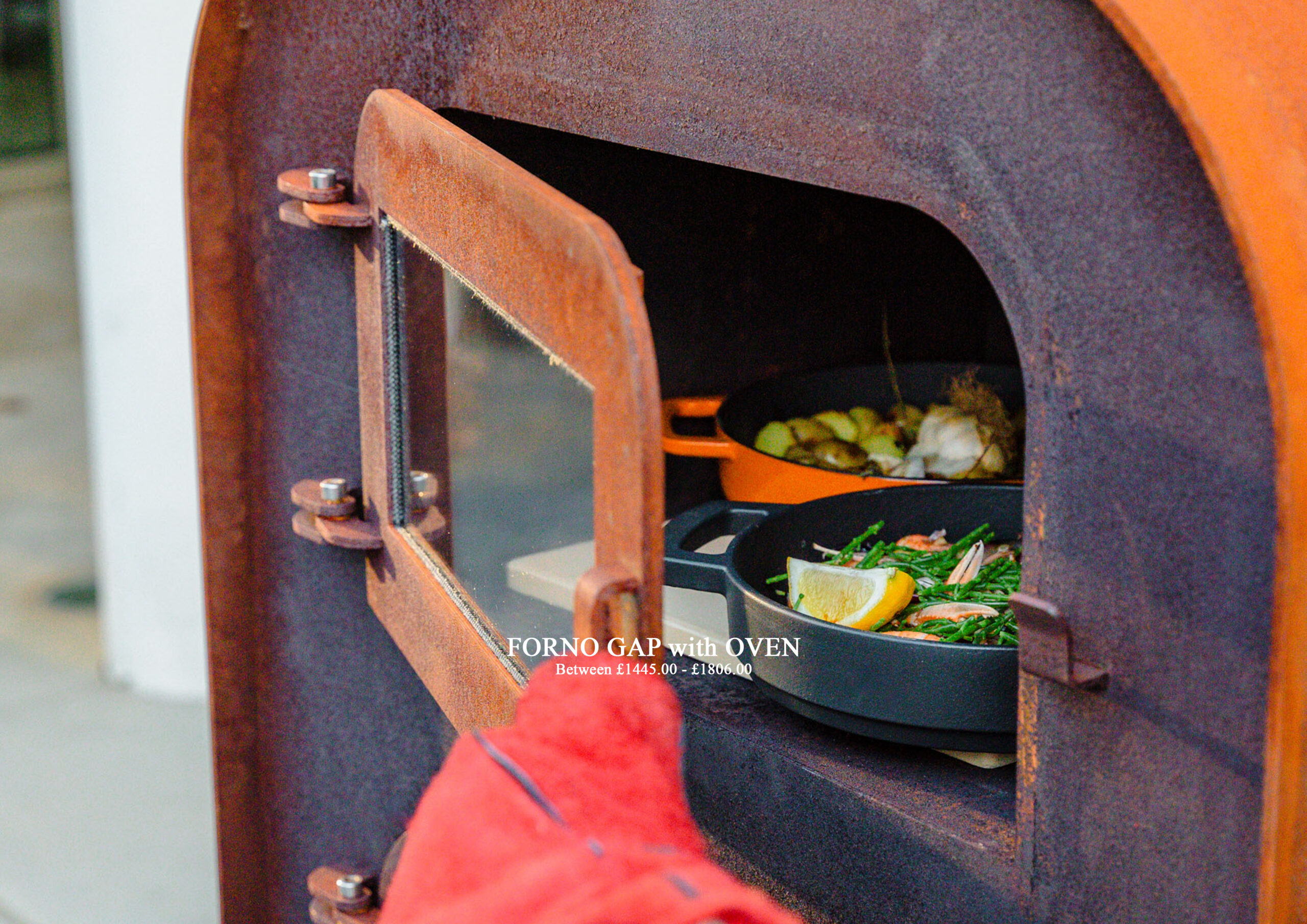 Close-up of a wood-fired oven door with a pan of vegetables (lemon wedges, greens) inside, showing a rusty orange exterior and a red cloth in the foreground. Screen text reads 'FORNO GAP with OVEN' and price range underneath.